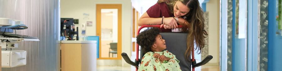 Health care worker pushes child patient down hospital hallway in wheelchair.